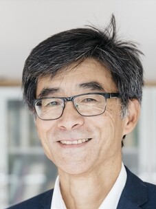 Herr Tjeng mit Brille, grauem Haar und Anzug, lächelnd.